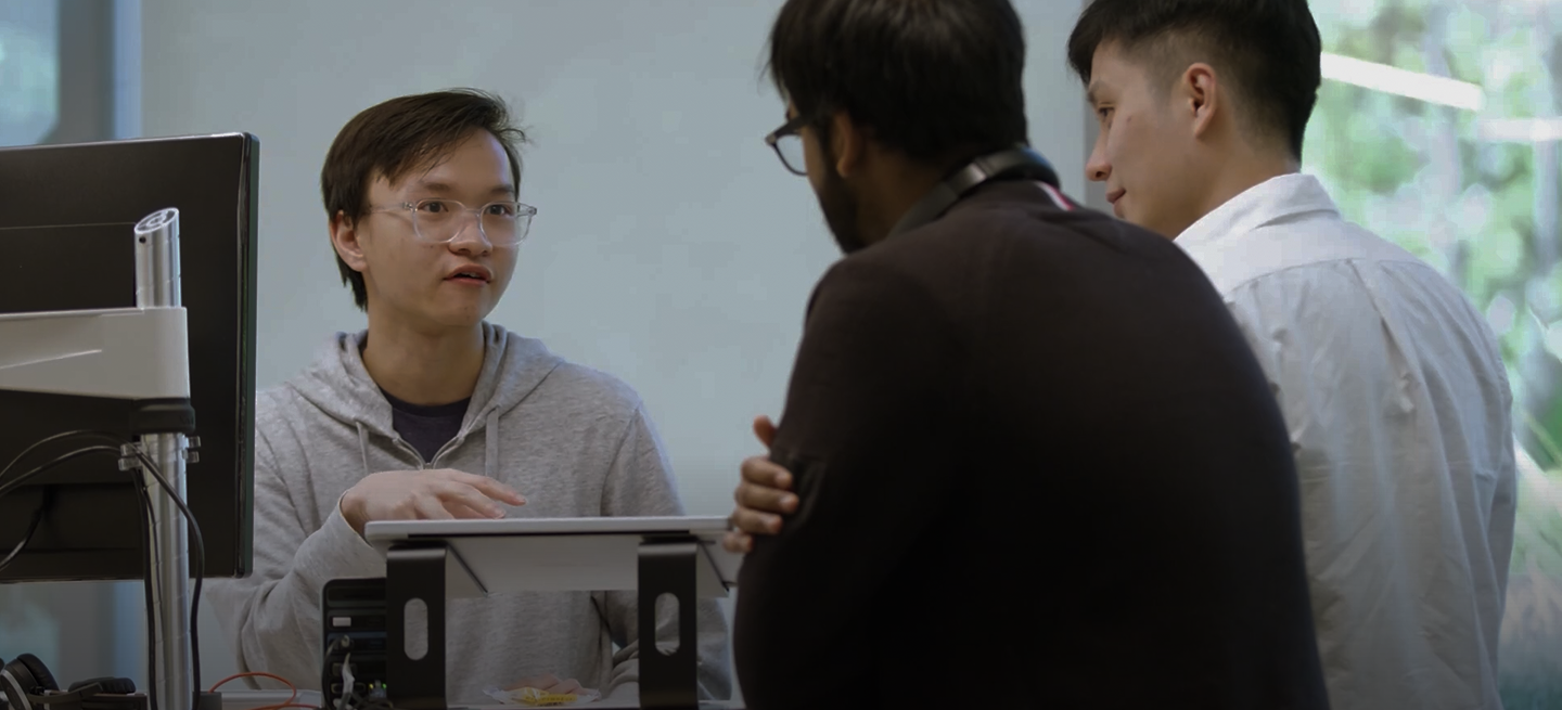 Background image of Three colleagues collaborating in an office, discussing work around a desk with a computer and documents